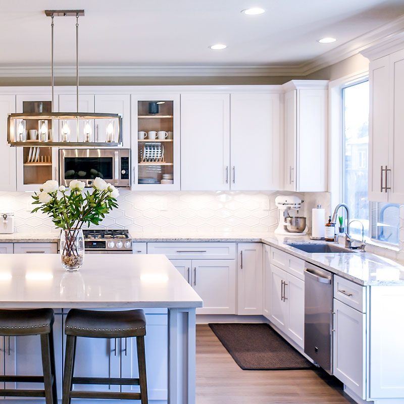A stylish and clean kitchen organized with plates and cups in the cupboards. A vase of flowers is centered on the island counter and a kitchen aid mixer sits on the counter near the sink. Daylight shines through the window above the sink.